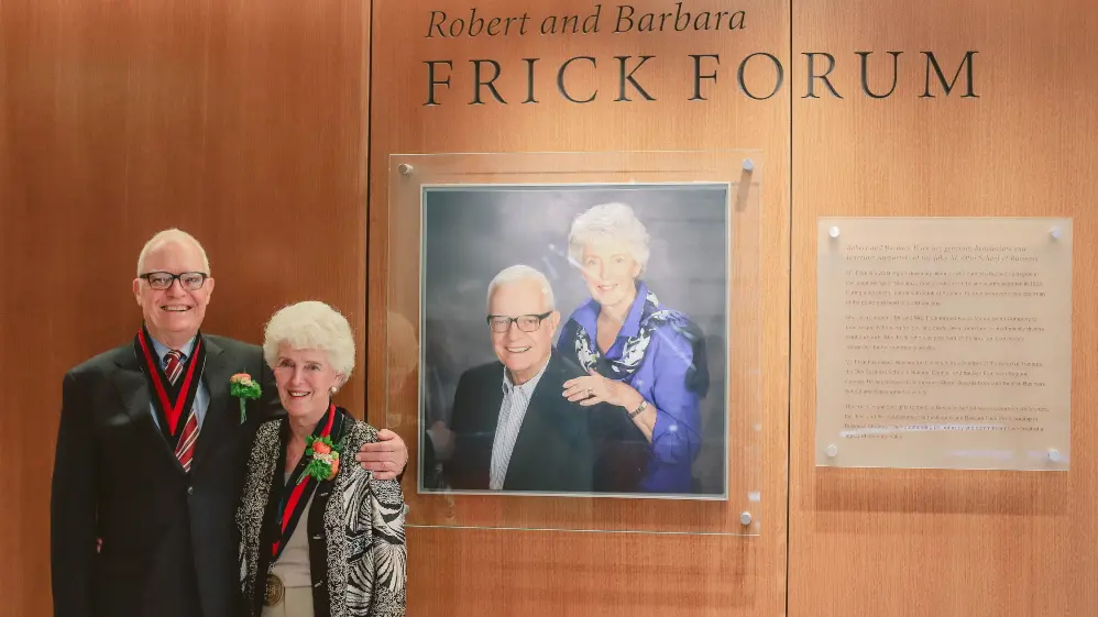 Robert and Barbara Frick standing in front of their portrait at the Frick Forum dedication wall