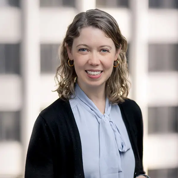 Professional headshot of Emily Koons Jae, a smiling woman with short wavy blonde hair wearing a light blue blouse with a bow and black cardigan, standing in front of a blurred office window background