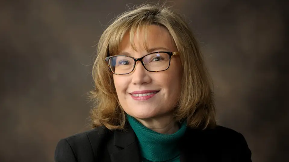 Professional headshot of Meg Cline wearing glasses, a black blazer, and green turtleneck against a studio background
