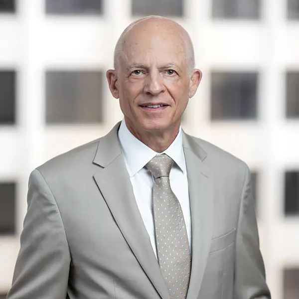 Professional headshot of Michael B. Poliakoff, Ph.D., a senior businessman in a beige suit and tie, standing in front of a blurred office window background