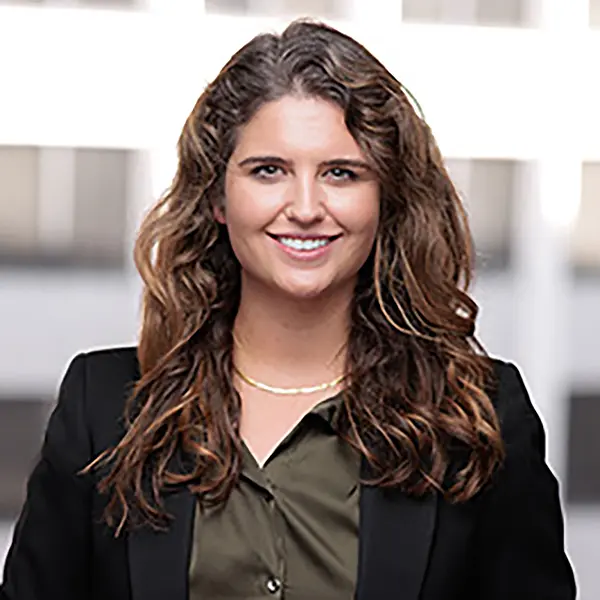 Professional headshot of Rebecca Richards, a smiling woman with long wavy brown hair, wearing a black blazer over an olive green blouse and gold necklace, standing in front of a blurred office background