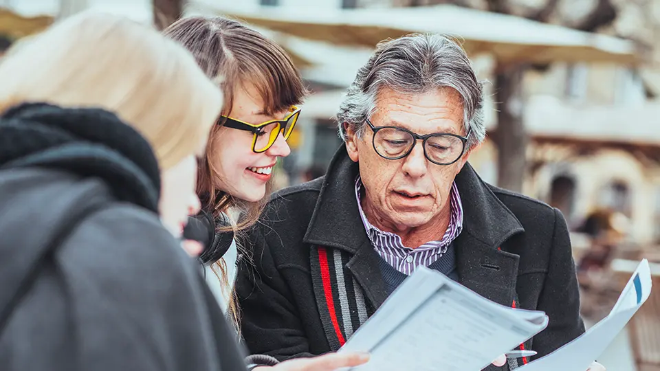 An older man is explaining documents to two young women outdoors