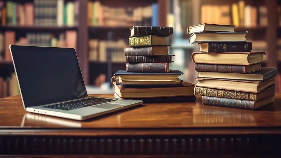 Laptop and stacks of academic books on a desk inside a university library study area