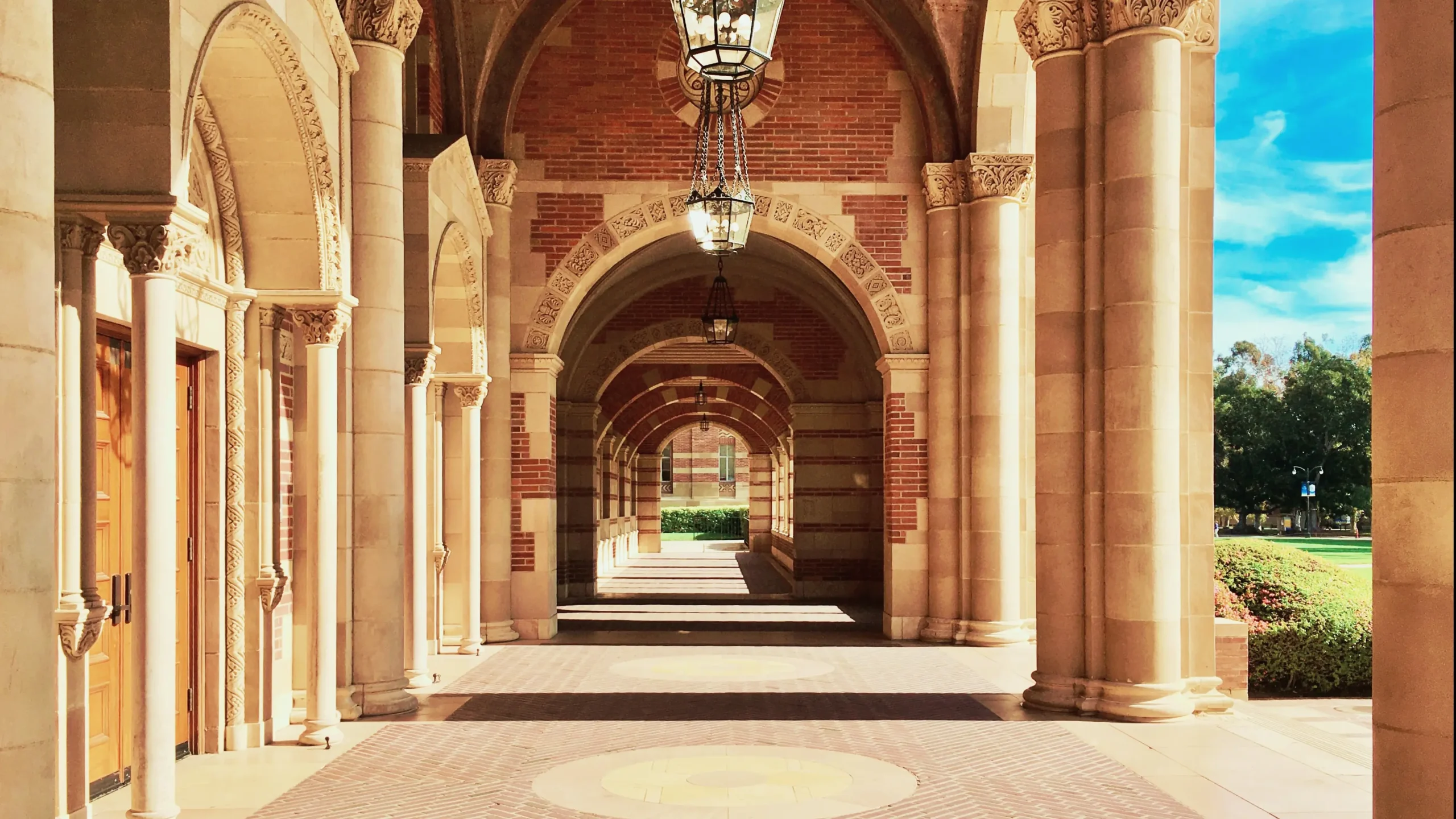 Elegant university campus corridor with stone columns, brick arches, and hanging lanterns in bright sunlight