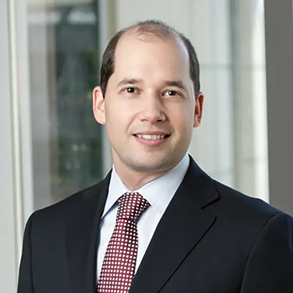 Professional headshot of Andras Kosaras, Esq., a smiling man with short dark hair wearing a black suit, light blue shirt, and red patterned tie, standing in front of a modern glass office background